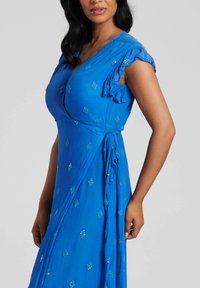 Abito blu con maniche a falda, cucitura verticale e design avvolgente. Presenta un tessuto testurizzato e piccole decorazioni.