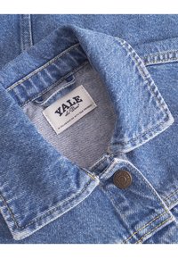 Col de manteau en denim bleu avec des coutures jaunes, présentant un bouton en métal rond et une étiquette blanche avec le texte "YALE" et un texte de collaboration.