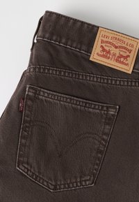 Poche arrière d’un jean en denim marron foncé avec un patch en cuir Levi Strauss & Co. et une étiquette en tissu rouge Levi's sur la couture latérale.