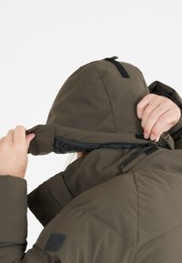 Grønne pufferjakke med en aftagelig hætte, der har lynlås lukning og velcro detaljer. Materialet fremstår glat og isoleret.