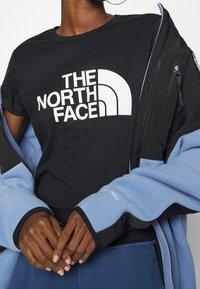 Person som bär en svart The North Face-t-shirt och en blå jacka som är delvis av, med händerna sammanfogade framför sig, beskuren vid axlarna och midjan.