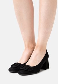 Escarpins en daim noir avec une boucle carrée sur le bout, dotés d'un talon épais pour le maintien et d'un bout arrondi.