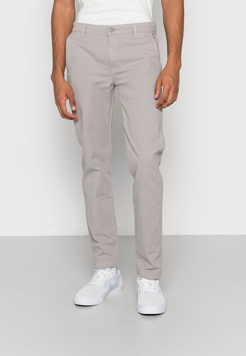 Chinos gris clair en mélange de coton, avec jambes droites, poches latérales et fermeture à bouton à l'avant. Portés avec des baskets blanches.