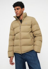 Khaki puffer takki, jossa on korkea kaulus, vetoketjusuljenta ja kaksi sivutaskua. Kangas on sileä, jossa on vartalonmyötäinen design ja hienovaraista logodetailia.