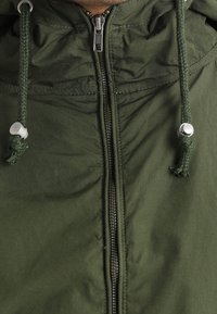 Zielona kurtka na zamek z kapturem, wyposażona w sznurki ściągające, metalowy zamek błyskawiczny i teksturowaną tkaninę. Posiada dwa widoczne przednie kieszenie.