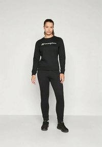 Moteris stovi vilkėdama juodą Champion aprangą - džemperį, leggingus ir juodus sportbačius prieš paprastą šviesią fono.