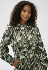 Rochie camasă cu mânecă lungă, în imprimeu abstract verde, crem și negru. Dispune de o zonă cu nasturi în față, guler și un Curea legată în talie. Textură de țesătură netedă.