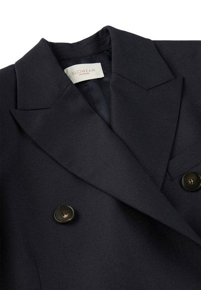 Blazer blu navy con due bottoni, bavero a punta e una texture morbida. L'etichetta recita "SLOWEAR." Realizzato in tessuto resistente.