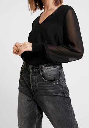 Mujer con blusa negra transparente de manga larga con puños fruncidos y pantalones vaqueros negros desgastados de talle alto, manos suavemente entrelazadas a la altura de la cintura.