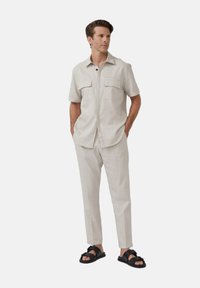 Chemise courte à manches courtes de couleur beige clair avec deux poches avant, assortie à un pantalon fuselé et des sandales à glisser noires. Texture lisse et tissée.