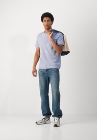 Tricou striped albastru, blugi de denim deschis și sneakers gri. Poartă o geantă tote bej și marin. Ținută casual cu o croială relaxată.