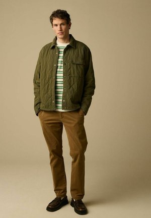 Jonge man staat met handen in de zakken, draagt een olijfgroene gewatteerde jas, een wit-groen gestreept shirt, bruine ribfluwelen broek en donkerbruine schoenen.