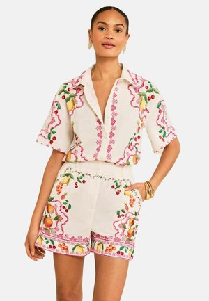 Vrouw draagt een bijpassende set van een crèmekleurig shirt met korte mouwen en korte broek met kleurrijke fruit- en bloemenprints, gouden oorbellen en armbanden.