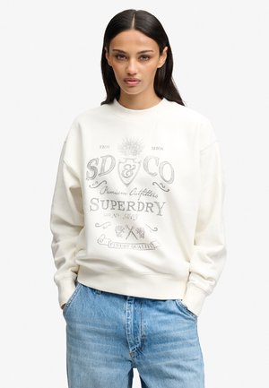 Kobieta z długimi ciemnymi włosami nosi biały sweter z szarym logo Superdry w stylu vintage oraz jasnoniebieskie dżinsy, ręce w kieszeniach, neutralne tło.