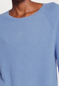 Pull bleu clair en maille avec un col rond et un tissu texturé, manches raglan et détails de couture subtils.