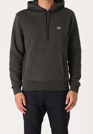 Man met een donkergroene hoodie met voorzak en klein krokodillenlogo op de borst, gecombineerd met zwarte broek.
