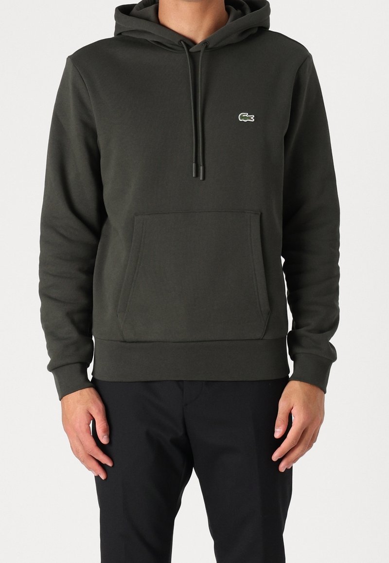 Homme portant un sweat à capuche vert foncé avec poche avant et petit logo crocodile sur la poitrine, associé à un pantalon noir.