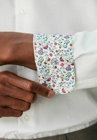 Bela srajca s cvetličnim potiskom na manšetah, ki vključuje majhne rdeče, vijolične in modre cvetove na belem ozadju. Teksturiran material.