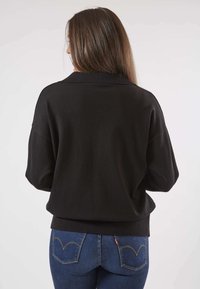Pull en laine noire avec une coupe décontractée, doté d'épaules basses et d'un ourlet côtelé, associé à un jean bleu mettant en valeur un détail de couture sur les poches arrière.