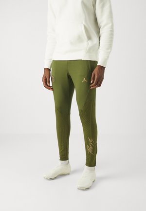 Homme portant un pantalon de sport moulant vert olive avec les logos Paris et Jordan, un sweat à capuche blanc, des crampons de football blancs, debout devant un fond uni.