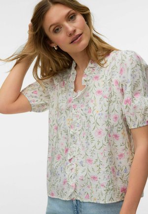 Kobieta o jasnobrązowych włosach, ubrana w białą kwiecistą bluzkę z falbanianym kołnierzem i krótkimi bufkowymi rękawami, zestawioną z jasnoniebieskimi dżinsami.