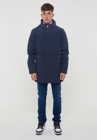 Giacca impermeabile blu navy con cappuccio, zip frontale, tasche laterali e un design elegante. Abbinata a jeans blu e sneakers nere.