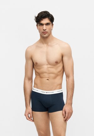 Mężczyzna bez koszulki z krótkimi ciemnymi włosami, ubrany w granatowe bokserki Tommy Hilfiger, stojący na jednolitym białym tle.