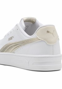 Zapatilla blanca con una franja dorada texturizada, parte superior de cuero suave, collar acolchado y suela de goma. Logotipo de PUMA en el lateral y el talón.