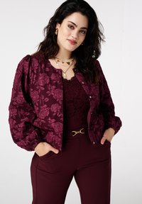 Bordeauxrode jas met bloemenprint en pofmouwen met metalen knopen, gedragen over een kanten top en gecombineerd met bijpassende broek, voorzien van een decoratieve riem.