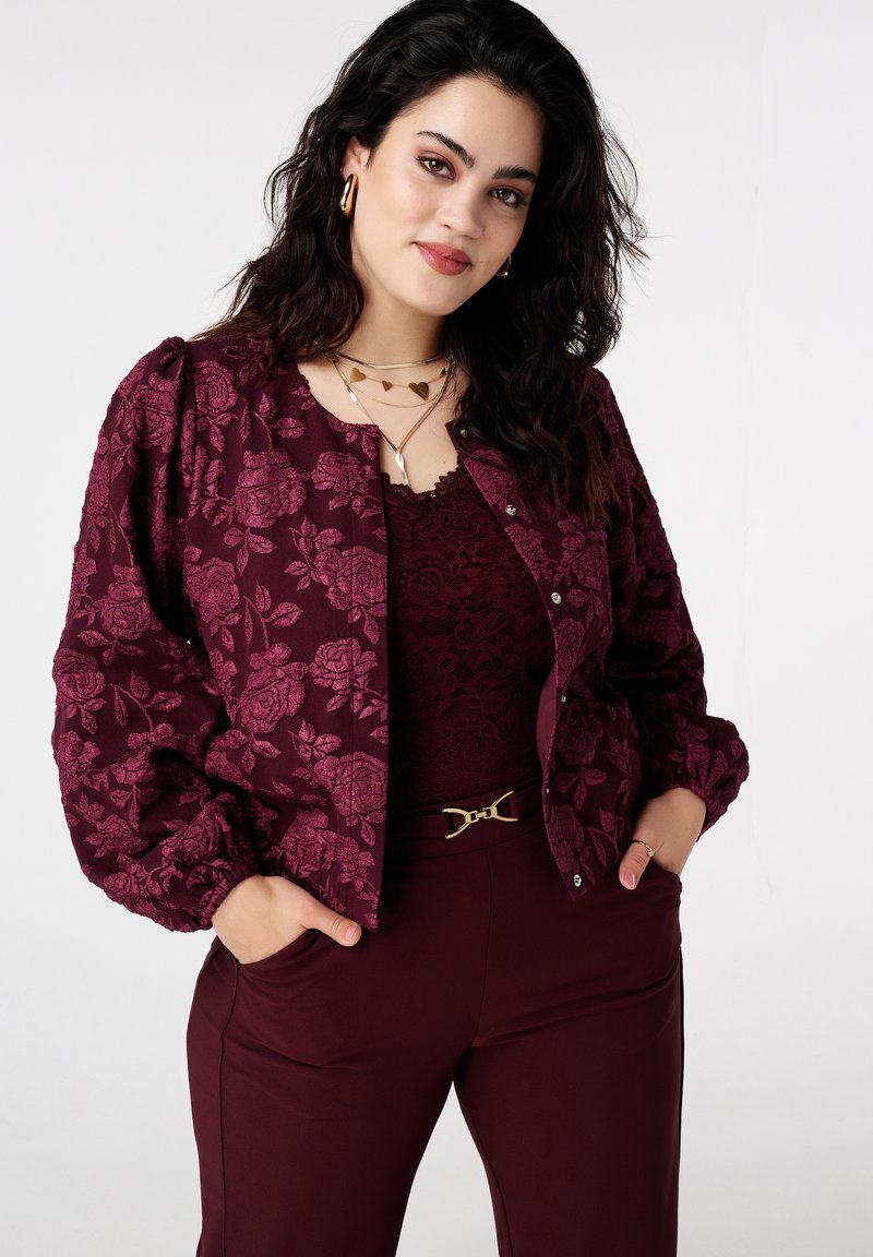 Bordeauxrode jas met bloemenprint en pofmouwen met metalen knopen, gedragen over een kanten top en gecombineerd met bijpassende broek, voorzien van een decoratieve riem.