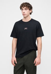 T-shirt de algodão preta com um pequeno emblema bordado no peito. A camisola tem um corte descontraído, mangas curtas e decote redondo.