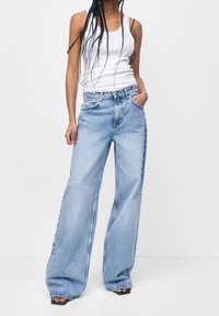 Jeans de mezclilla de pierna ancha en azul claro con cintura alta, que presentan un diseño clásico de cinco bolsillos y costuras visibles. Combinado con una camiseta sin mangas blanca.