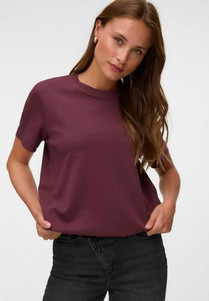 Kvinde med langt brunt hår iført en lilla kortærmet skjorte og sorte jeans, stående med hænderne i lommerne mod en neutral baggrund.