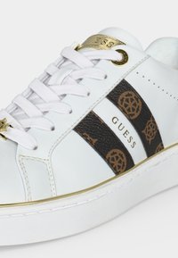 Baskets blanches Guess avec lacets blancs, bandes latérales marron à motif, accents dorés et logo sur le côté et la languette.