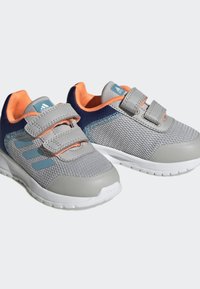 Baskets de sport grises avec tige en maille, accents oranges et straps en velcro. Présentent un renfort au niveau de l'orteil et une semelle en caoutchouc blanche.