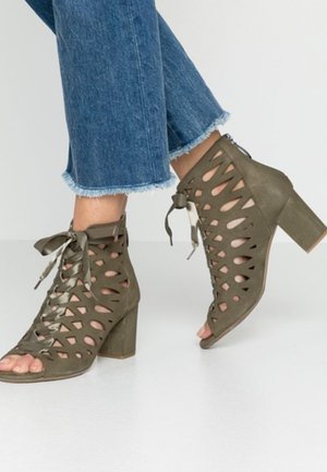 Person trägt blau ausgefranste Jeans und olivgrüne Cut-Out-Schnürsandalen mit Blockabsatz, Zehen sichtbar, steht auf einer hellen Oberfläche.
