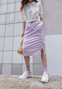 Sudadera blanca recortada con logo de Nike, falda morada clara fruncida, zapatillas pastel y bolso marrón texturizado. Calcetines morados con bordes ondulados.