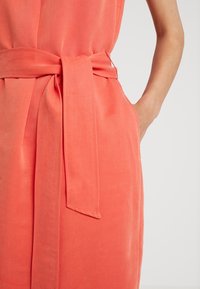 Robe corail à texture lisse, avec une ceinture à la taille et des poches latérales, conçue avec une coupe décontractée.