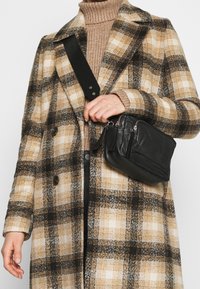 Svart läder crossbody-väska med flera zippered fack, i kombination med en rutig, beige och svart ullrock med stora knappar.