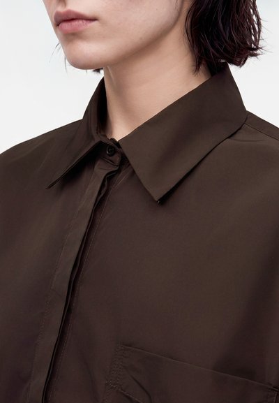 Gros plan d'une personne portant une chemise marron foncé à col, avec patte de boutonnage et poche poitrine, sur un fond clair uni.