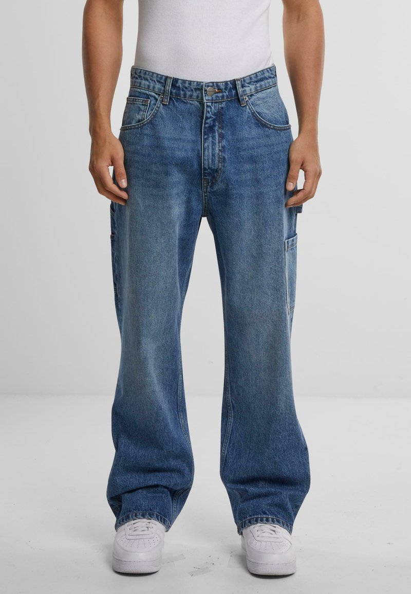 Jeans de denim azul de pierna ancha con un lavado desvanecido, bolsillos delanteros y un bolsillo cargo lateral. Costuras acolchadas y cierre de botón en la cintura.