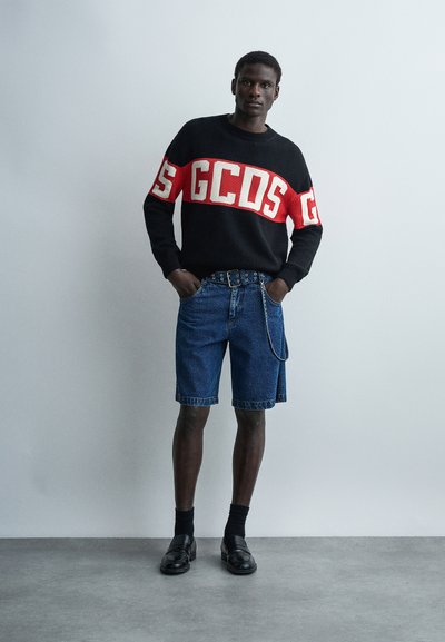 Άνδρας που στέκεται με τα χέρια στις τσέπες, φοράει μαύρο και κόκκινο πουλόβερ GCDS, τζιν σορτς με ζώνη και αλυσίδα, μαύρες κάλτσες και μαύρα παπούτσια.