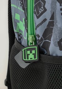 Mochila con un tejido gris con patrones geométricos negros y acentos verdes, cierre con cremallera y tirador de cremallera con temática de Minecraft en verde.