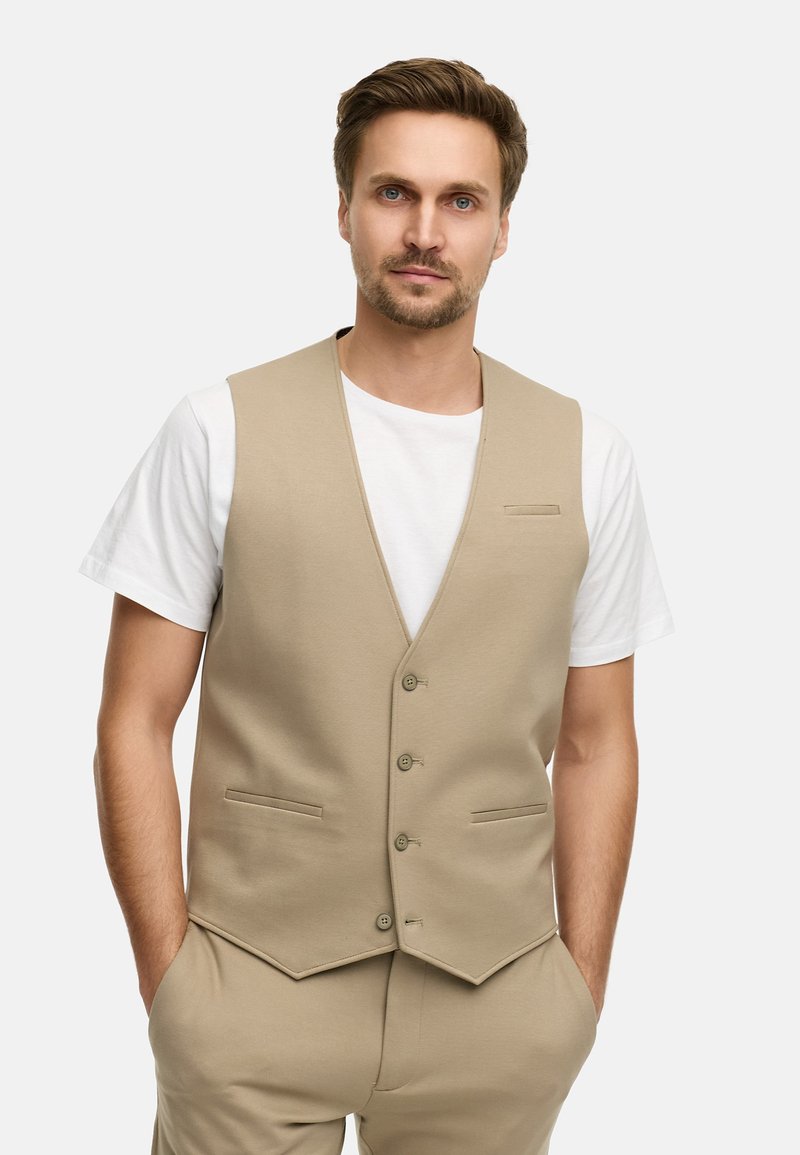 Beige vest laget av glatt stoff, med V-hals, fem knapper, to frontlommer og en litt avsmalnet kant. Brukt over en hvit skjorte.