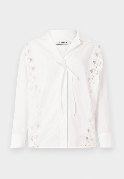 Hvit skjorte laget av bomull med omslag foran, blonde detaljer på sidene, lange ermer og krage. Knytes i livet.