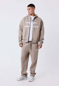 Beige hoodie met wit logo, gecombineerd met bijpassende joggers. Casual pasvorm met geribde manchetten en een verstelbare tailleband met trekkoord. Witte sneakers.