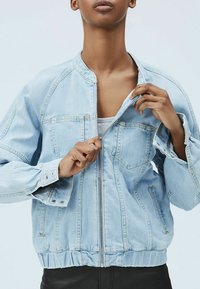 Chaqueta de mezclilla azul claro con cierre de cremallera en el frente, dobladillo acanalado y puños. Cuenta con dos bolsillos frontales y un corte relajado, ligeramente oversize.