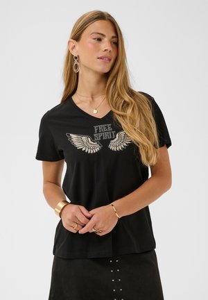 Kvinde med langt blond hår iført sort V-hals T-shirt med rhinstenvinger og teksten "FREE SPIRIT", guldsmykker og sort nederdel med nitter.