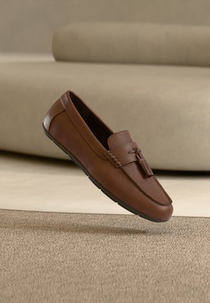Mocassin en cuir marron avec détail à pampilles, posé sur un tapis beige texturé devant un canapé beige clair.