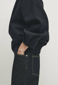 Navyblauwe oversized jacket met pofmouwen, voorzien van een Gathered zoom en zijzakken, gecombineerd met donkere spijkerbroeken en zichtbare stikdetails.